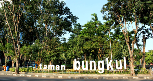 A vibrant view of Taman Bungkul Surabaya, featuring lush greenery, walking paths, and modern sculptures, with people relaxing, enjoying the park’s peaceful atmosphere, and a clear sky overhead.