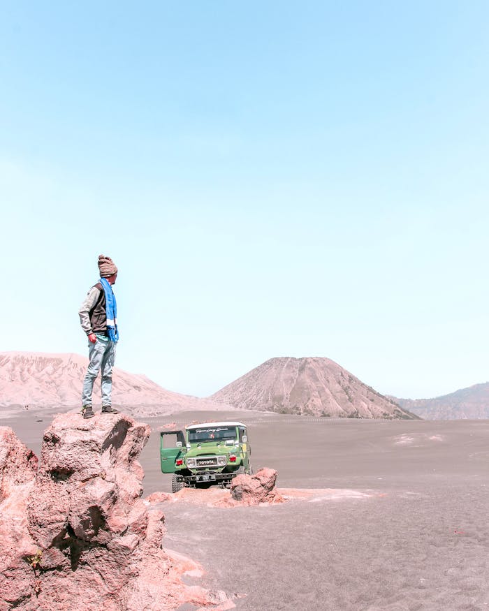 Man standing on rock in Bromo desert, exploring mountainous landscape with vehicle nearby.