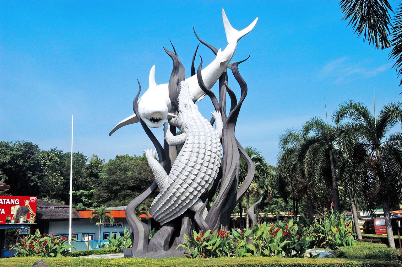 The iconic Surabaya Statue, depicting a fierce battle between a crocodile and a shark, symbolizing the city's strength and resilience, set against a bright blue sky and urban backdrop.