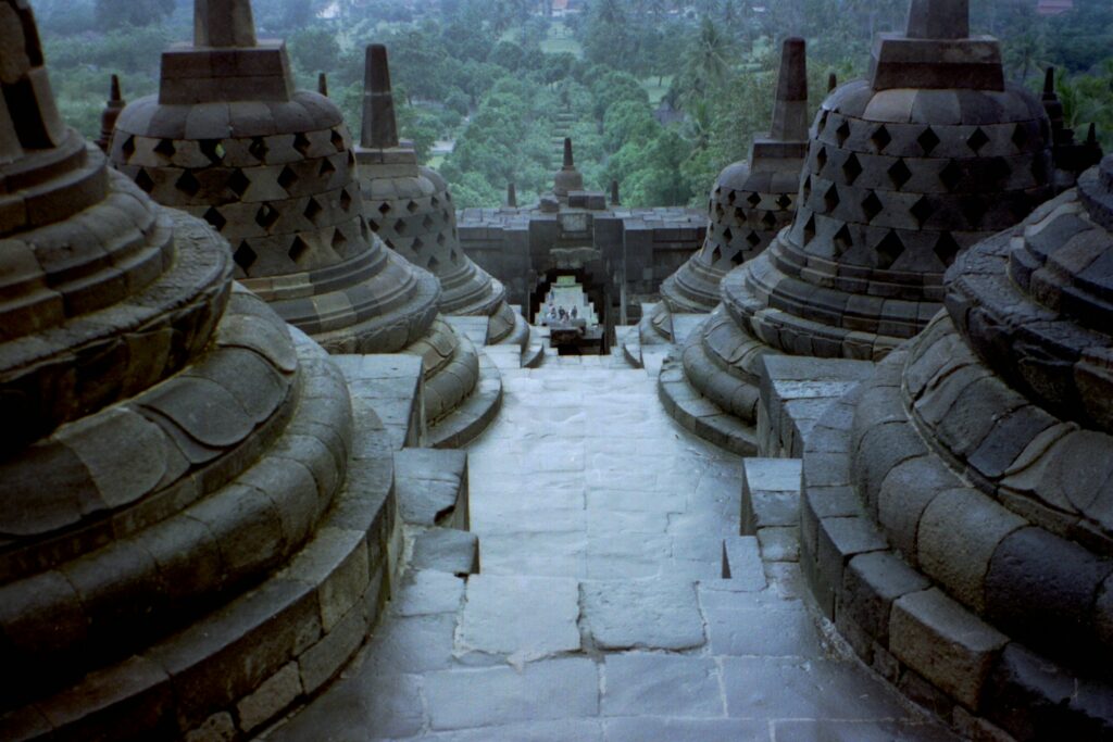 Bird's-eye view of Borobudur Temple, showcasing its tiered structure and intricate stone carvings, leading down to the surrounding green landscape.