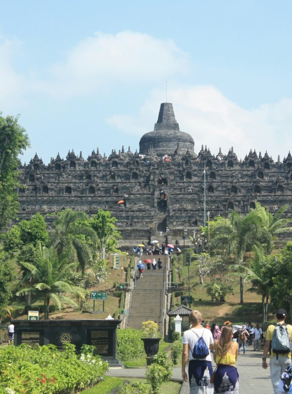 View from the base of Borobudur Temple, with tourists ascending the steps and exploring the monument's intricate carvings, surrounded by lush greenery.