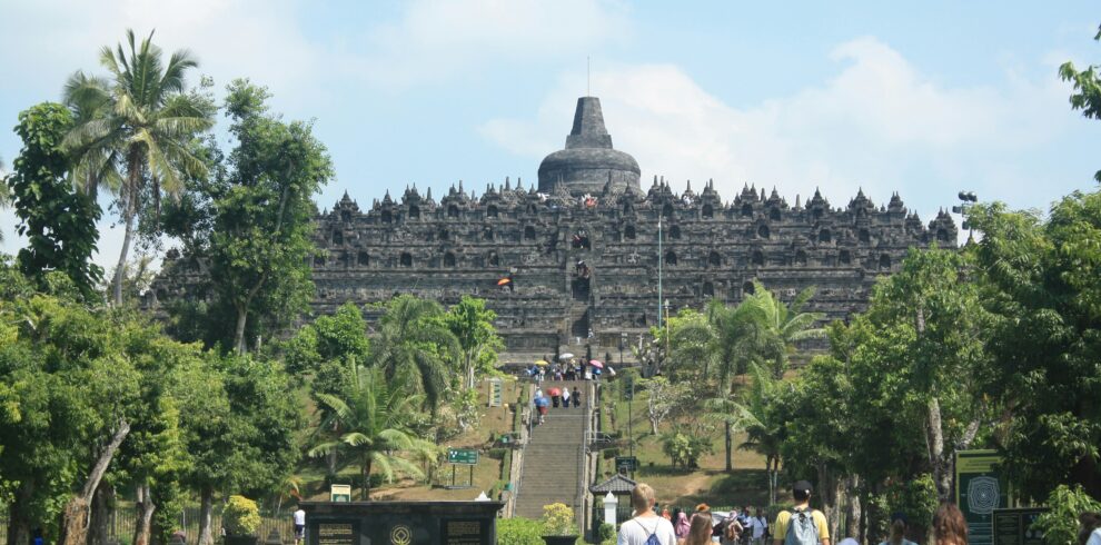 View from the base of Borobudur Temple, with tourists ascending the steps and exploring the monument's intricate carvings, surrounded by lush greenery.