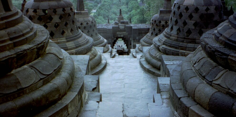 Bird's-eye view of Borobudur Temple, showcasing its tiered structure and intricate stone carvings, leading down to the surrounding green landscape.