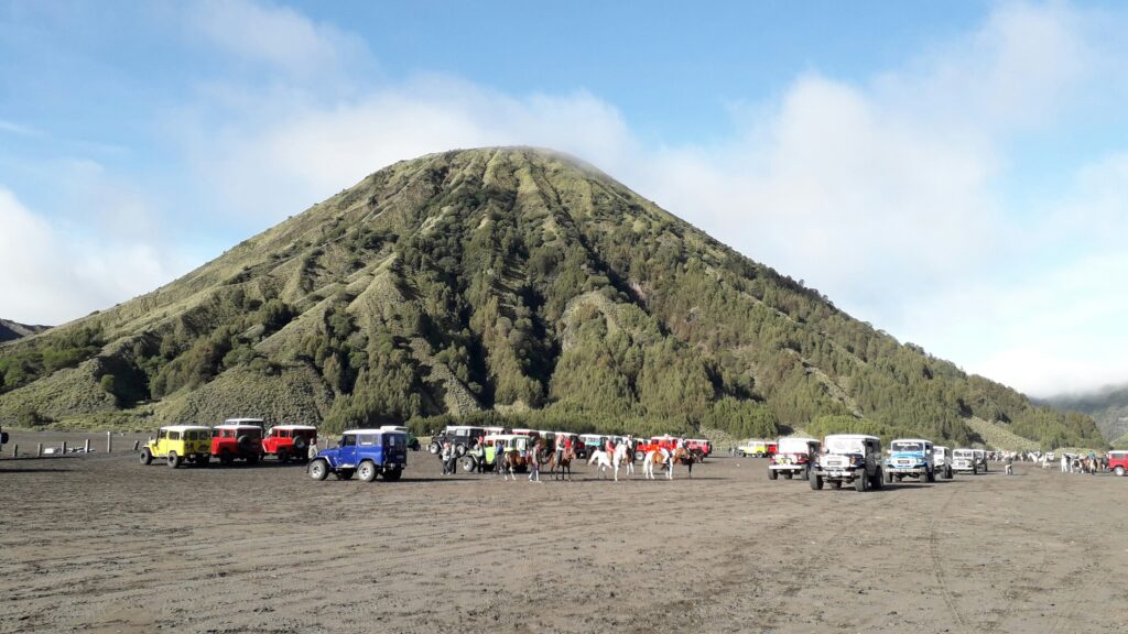 Tourists gathered at the base of Mount Bromo, exploring the vast volcanic plains with the iconic peak in the background, under a clear blue sky.