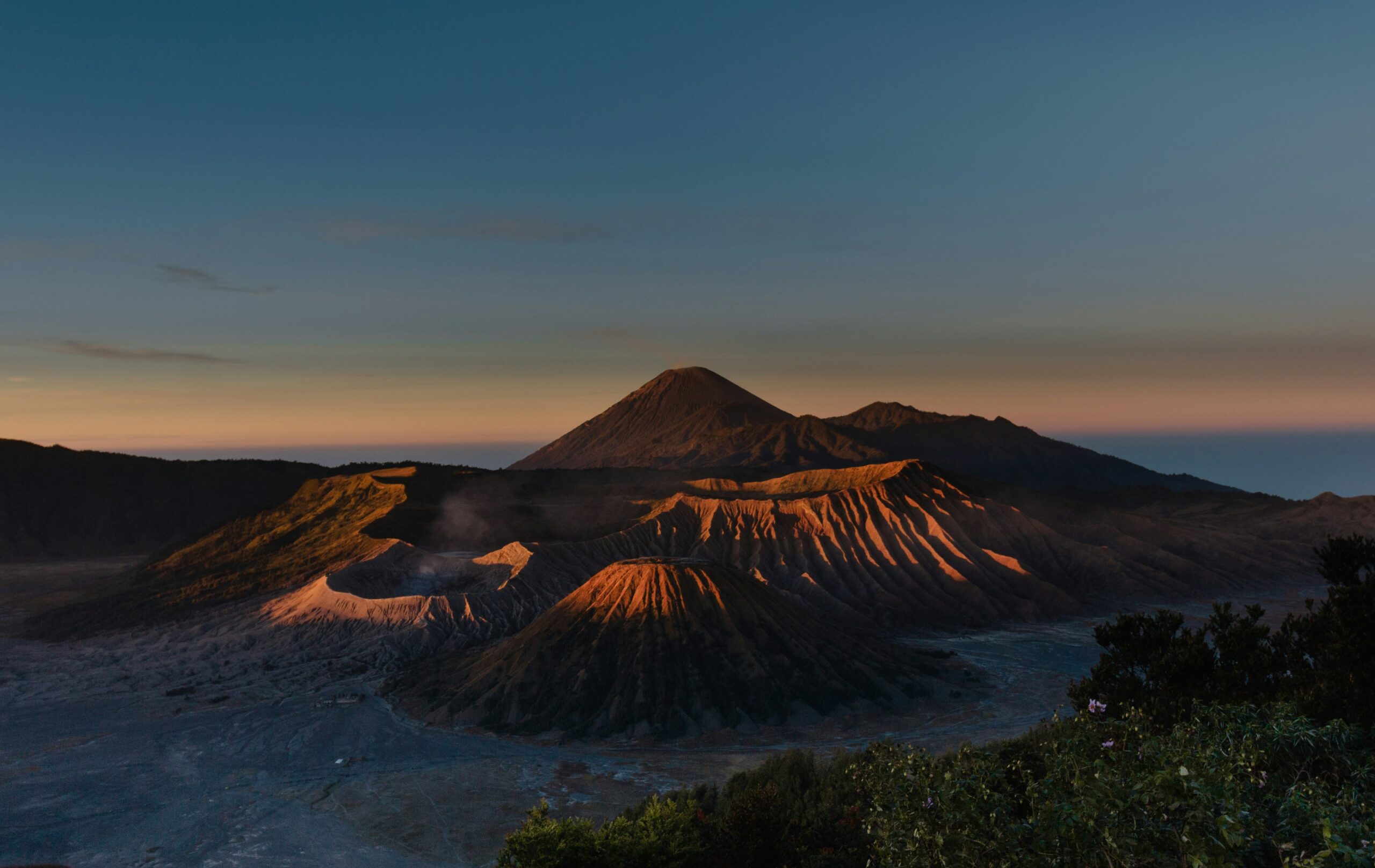 View of Mount Bromo from its base, showcasing the vast plains leading up to the towering volcanic peak, under a clear sky and surrounded by rugged terrain.