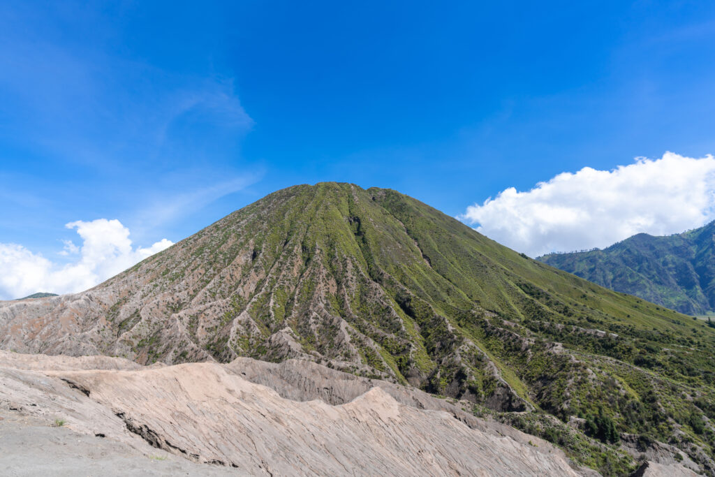 View of Mount Bromo from its base, showcasing the vast plains leading up to the towering volcanic peak, under a clear sky and surrounded by rugged terrain.