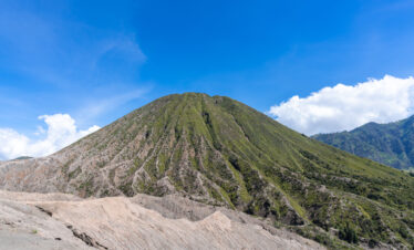 View of Mount Bromo from its base, showcasing the vast plains leading up to the towering volcanic peak, under a clear sky and surrounded by rugged terrain.
