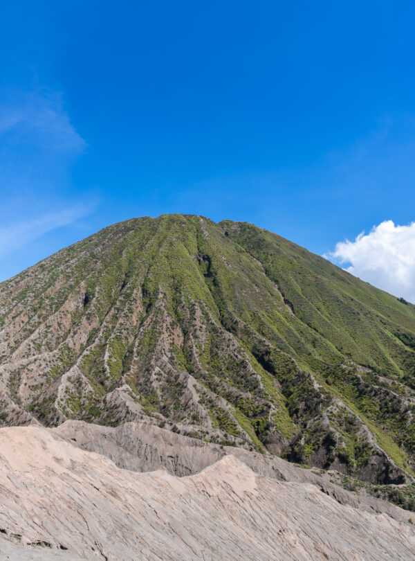 View of Mount Bromo from its base, showcasing the vast plains leading up to the towering volcanic peak, under a clear sky and surrounded by rugged terrain.