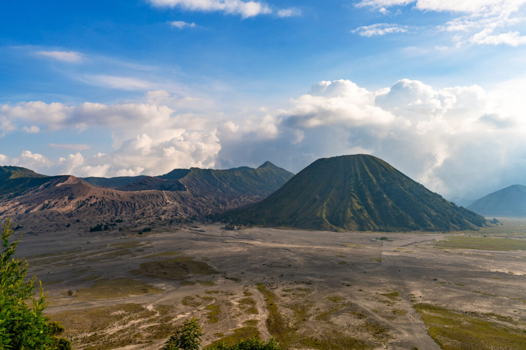 Clear view of Mount Bromo, with its rugged volcanic peak standing tall against a bright blue sky dotted with a few clouds, highlighting the surrounding barren landscape.