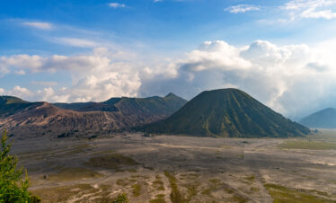 Clear view of Mount Bromo, with its rugged volcanic peak standing tall against a bright blue sky dotted with a few clouds, highlighting the surrounding barren landscape.