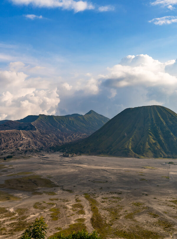 Clear view of Mount Bromo, with its rugged volcanic peak standing tall against a bright blue sky dotted with a few clouds, highlighting the surrounding barren landscape.