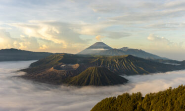 View of Mount Bromo with its mist-covered base, surrounded by a vast plain and a clear sky with a few clouds, creating a dramatic yet serene atmosphere.