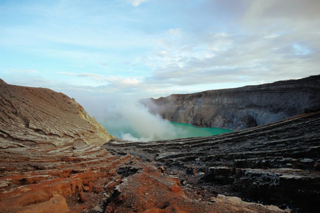Striking view of Kawah Ijen with its vibrant teal-colored acidic lake, contrasting against the orange rim of the crater, surrounded by rocky terrain and a clear sky.