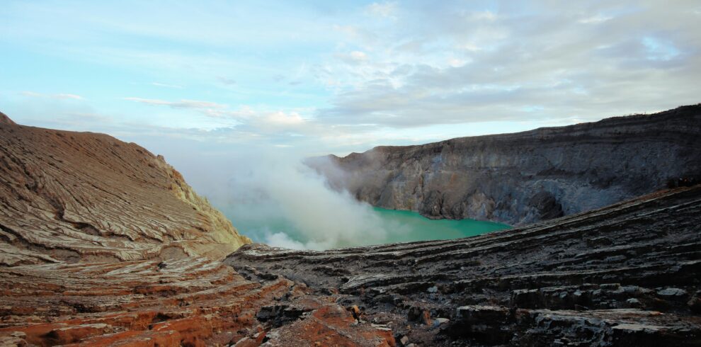 Striking view of Kawah Ijen with its vibrant teal-colored acidic lake, contrasting against the orange rim of the crater, surrounded by rocky terrain and a clear sky.