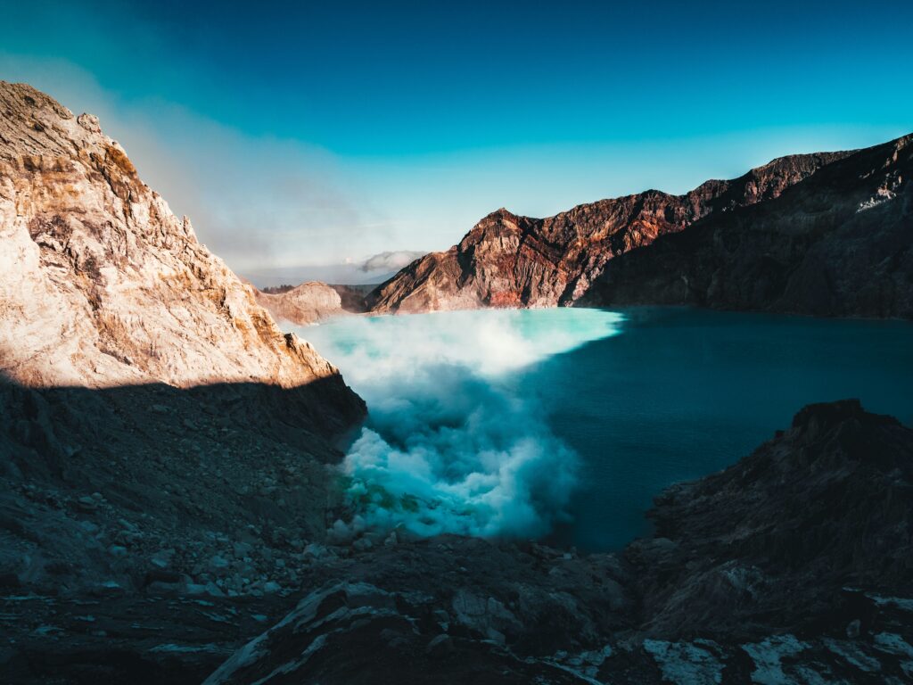 Panoramic view of Kawah Ijen, showcasing its vibrant teal-colored acidic lake surrounded by rugged volcanic terrain and a clear sky.