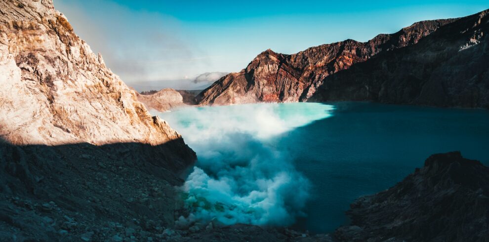 Panoramic view of Kawah Ijen, showcasing its vibrant teal-colored acidic lake surrounded by rugged volcanic terrain and a clear sky.