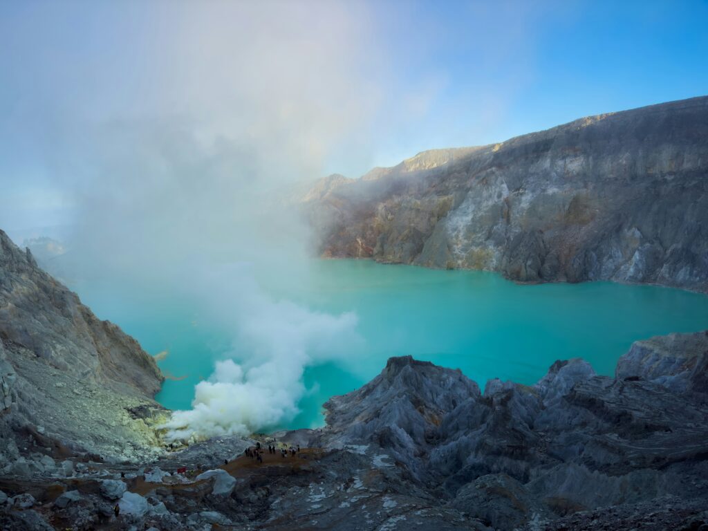 Tourists exploring the stunning Kawah Ijen, standing near the edge of the vibrant teal lake, with the surrounding volcanic landscape and smoke rising from the crater.