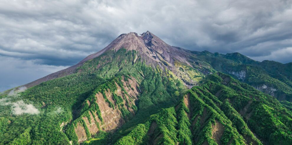 Mount Merapi standing tall against a clear blue sky, with its rugged volcanic slopes visible and the surrounding landscape bathed in sunlight.