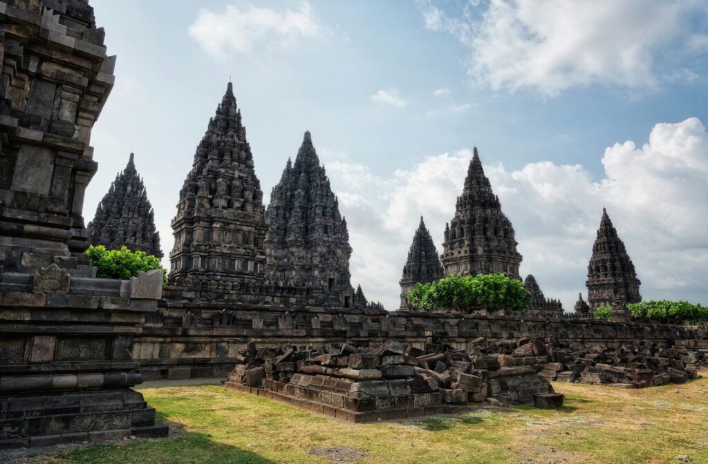 Prambanan Temple complex with towering ancient structures, some partially ruined, surrounded by scattered stones and a lush green landscape under a clear sky.