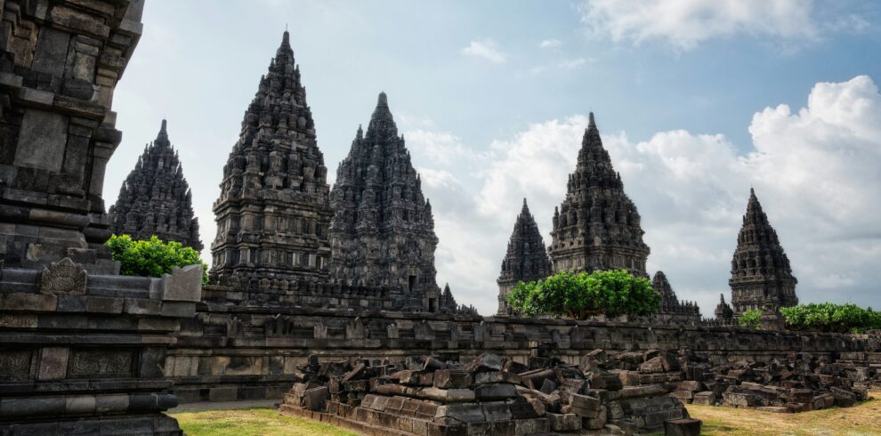 Prambanan Temple complex with towering ancient structures, some partially ruined, surrounded by scattered stones and a lush green landscape under a clear sky.