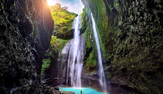 A man standing beneath the majestic Madakaripura Waterfall, with water cascading down the cliff and surrounding mist creating a dramatic and refreshing atmosphere.