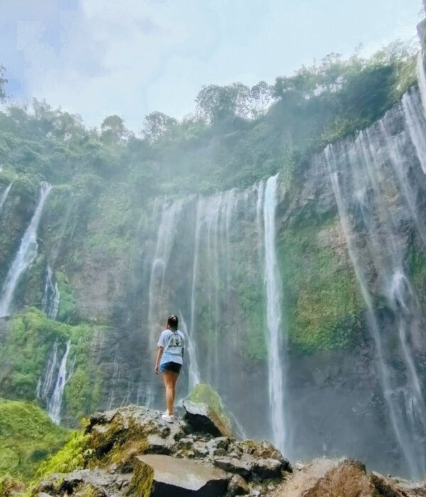 A woman standing beneath the powerful Tumpak Sewa Waterfall, with water droplets creating a fine mist around her as the cascading water crashes down, surrounded by lush greenery.