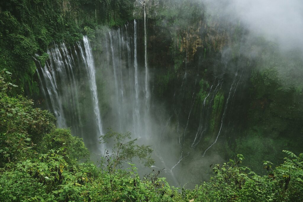 Overhead view of Tumpak Sewa Waterfall, with its powerful cascade partially hidden by clouds and surrounded by misty forests and rugged cliffs.