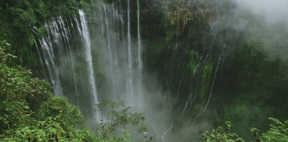 Overhead view of Tumpak Sewa Waterfall, with its powerful cascade partially hidden by clouds and surrounded by misty forests and rugged cliffs.