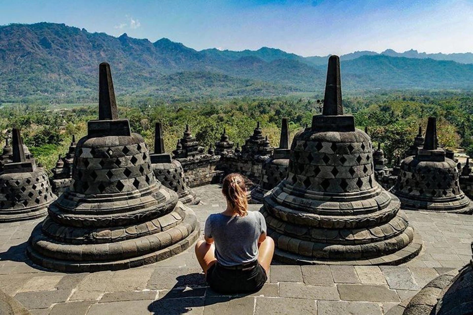 A person sitting on one side of the Borobudur temple, surrounded by the intricate stone carvings, with a clear blue sky and bright sunlight in the background.